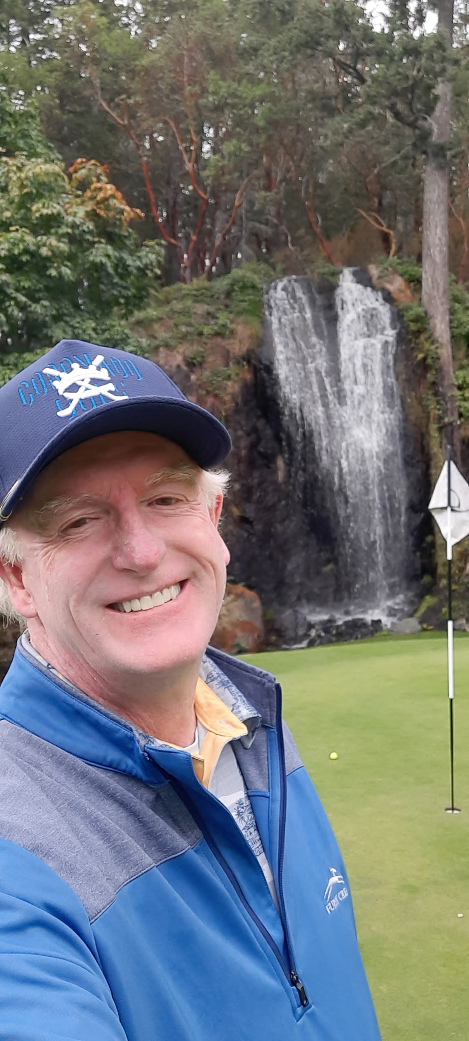 Man in a blue jacket and cap standing on a golf course with a waterfall in the background